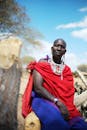 A bald man in traditional tribal clothing poses by a tree trunk outdoors.